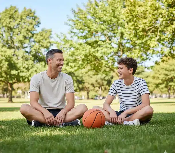 Father and son sitting on grass in a park with a basketball, smiling at each other