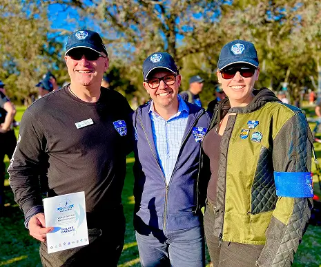 Three adults wearing caps and badges posing together at an outdoor event.