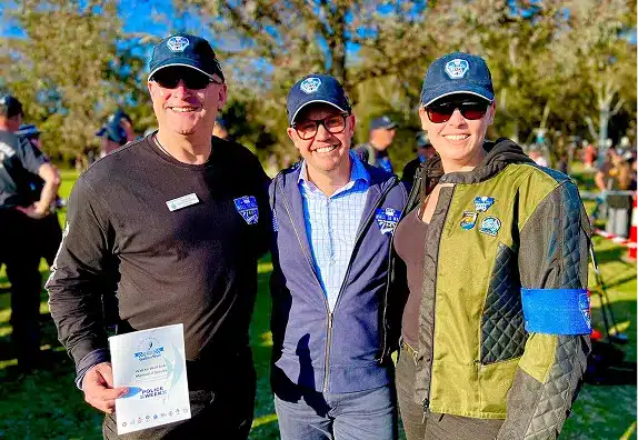 Three people wearing matching caps and badges, smiling together at an outdoor event.